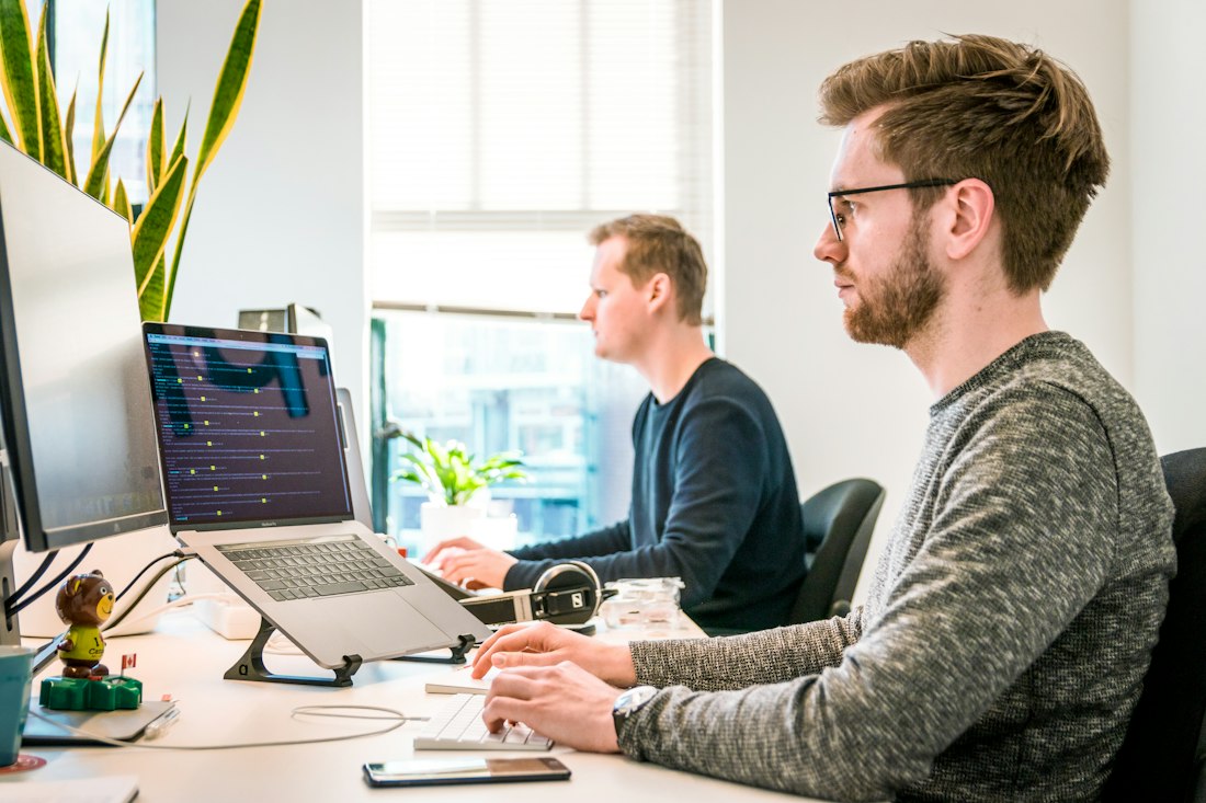 Two people working at desks with laptops and monitors in a bright office